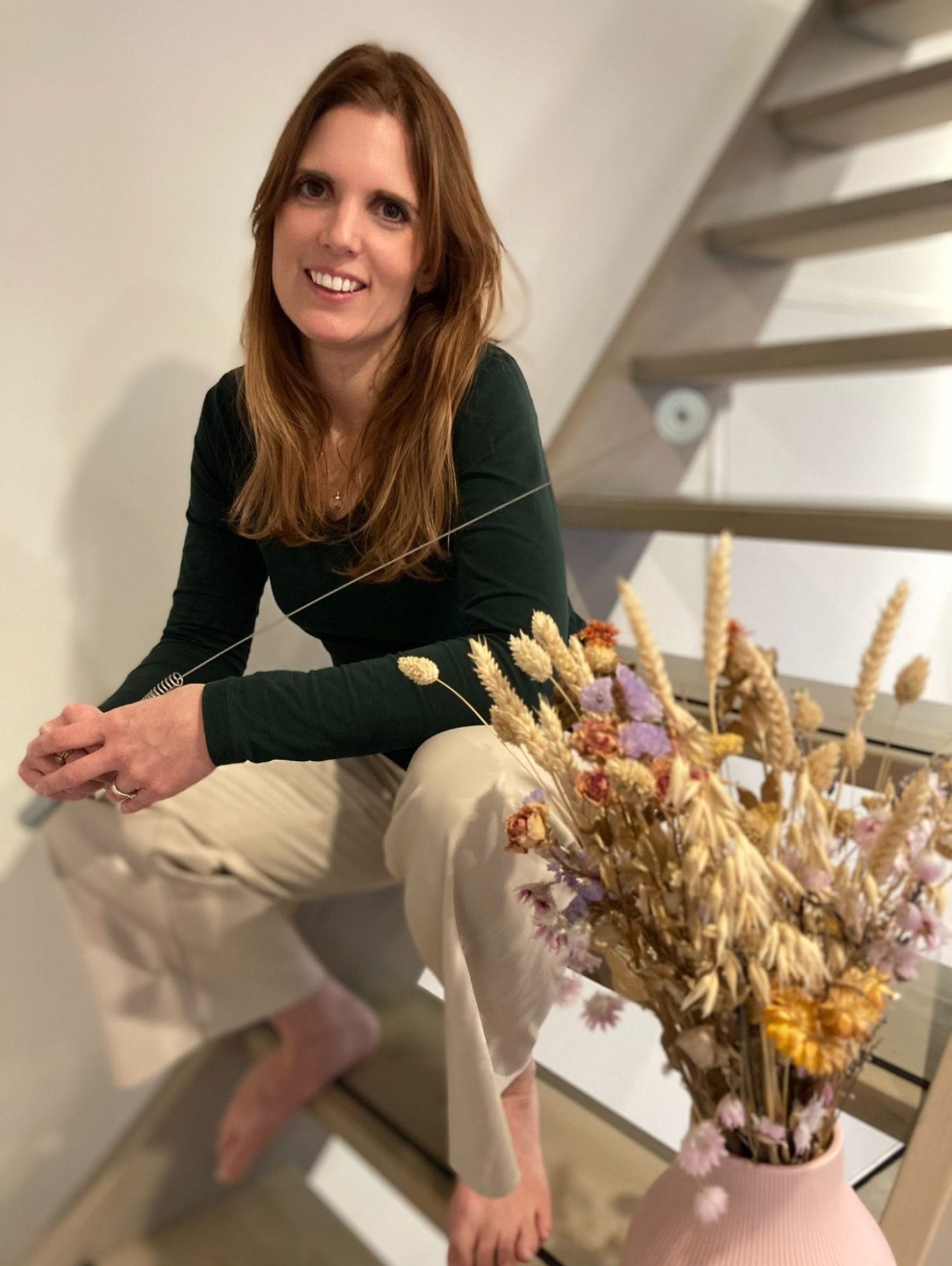 Vrouw met lange, kastanjebruine haren zittend op een trap, omringd door droogbloemen.