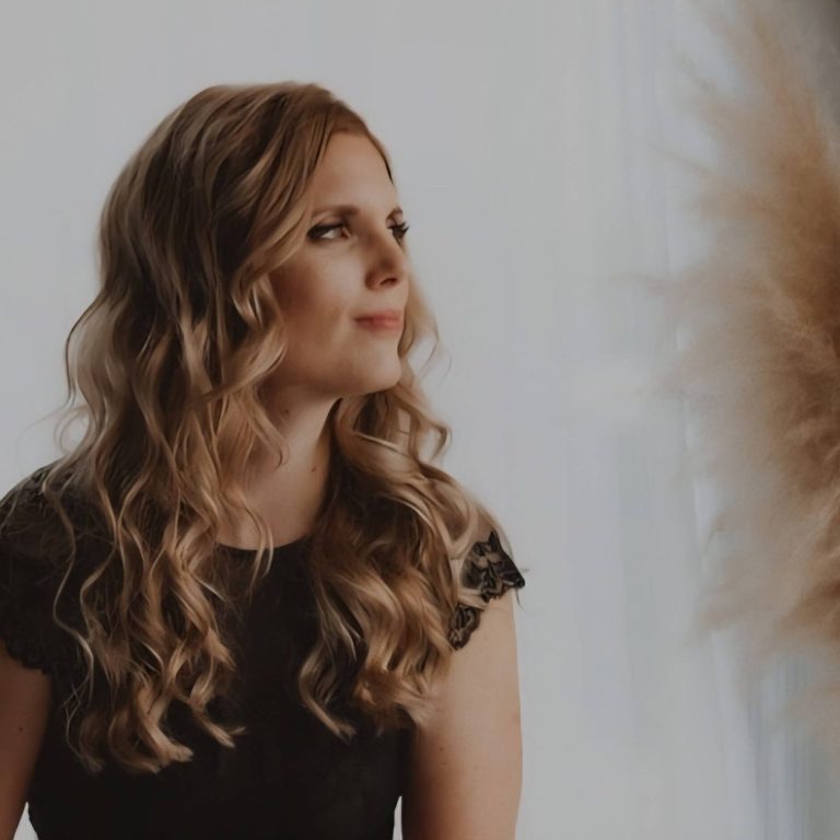 Vrouw met krullend haar in een donkere top, staande naast gedroogde bloemen. praktische hulp kinderwens online traject